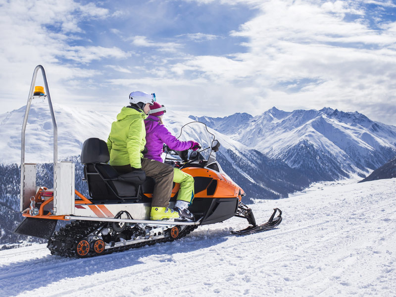 two people ride a snowmobile on a snowy path with mountain peaks in distance