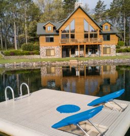 A large cabin sits on the shore of a private pond while a giant lounging raft floats on the surface of the pond.