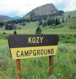 Wooden sign that reads "Kozy Campground" with green meadow and mountains in background