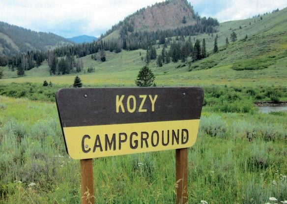 Wooden sign that reads "Kozy Campground" with green meadow and mountains in background