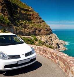 a white compact car parked at the edge of a bluff with turquoise blue sea and rising foothills in the background