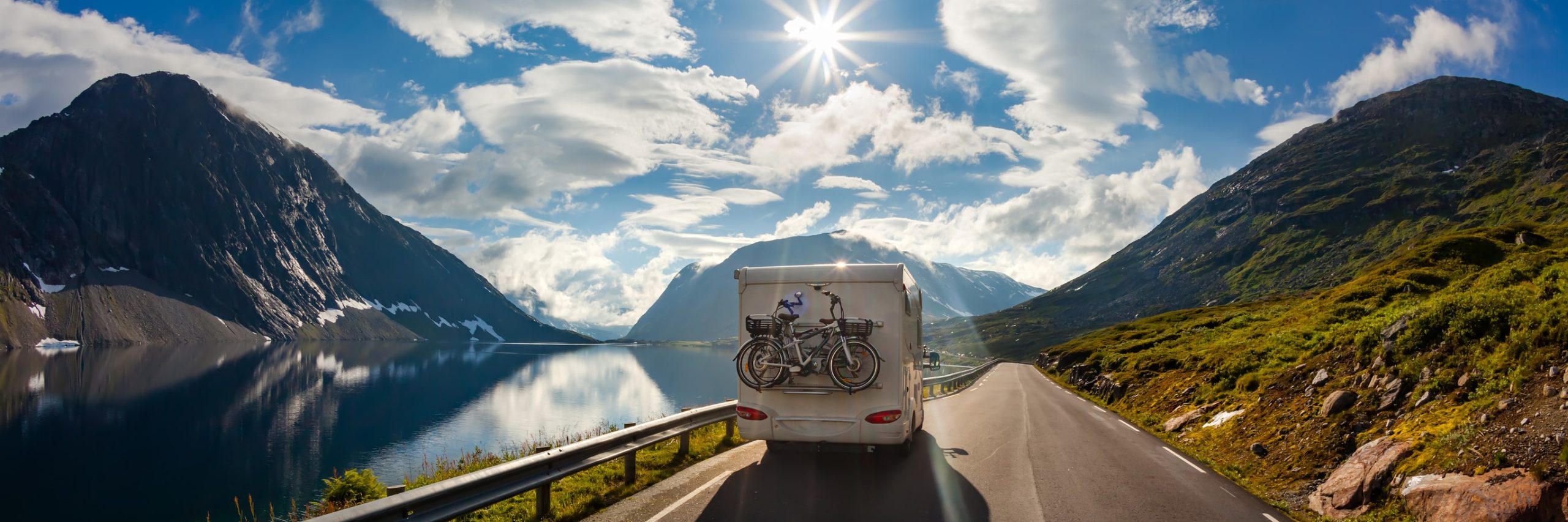 View from center of road of a motorhome driving away with a lake on the left, mountain on the right and sun directly overhead